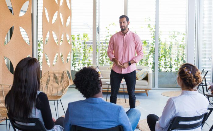 Man speaking to a group of three people in windowed room.